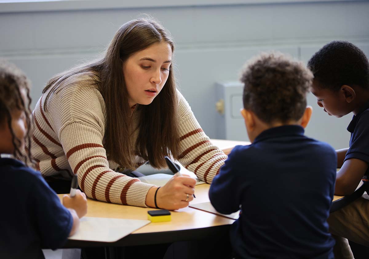 HSA Teacher smiles while kneeling beside a young student in a classroom setting.